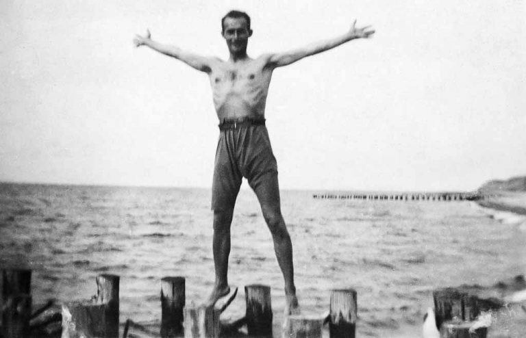 Ein junger Mann steht mit ausgebreiteten Armen auf den Bohlen der Ostsee.