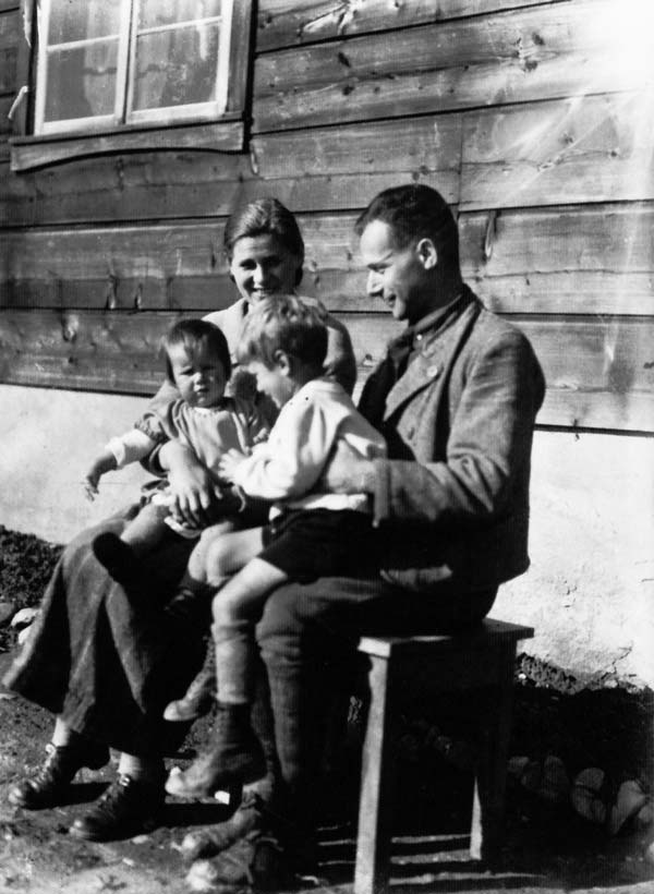 Friedrich und Else Wolf sitzen mit ihren kleinen Kindern Markus und Konrad vor ihrem Holzhaus.