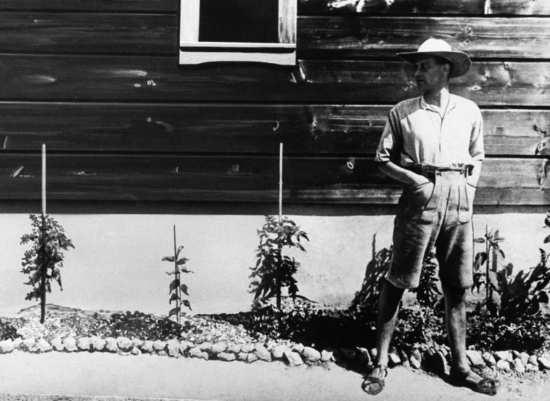 Friedrich&nbsp;Wolf steht mit Hut und Sandalen an einem Holzhaus mit bepflanzten Vorbeeten.